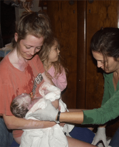 Woman with green shirt handing newborn baby to woman with an orange shirt. Child holding on to woman with orange shirt.